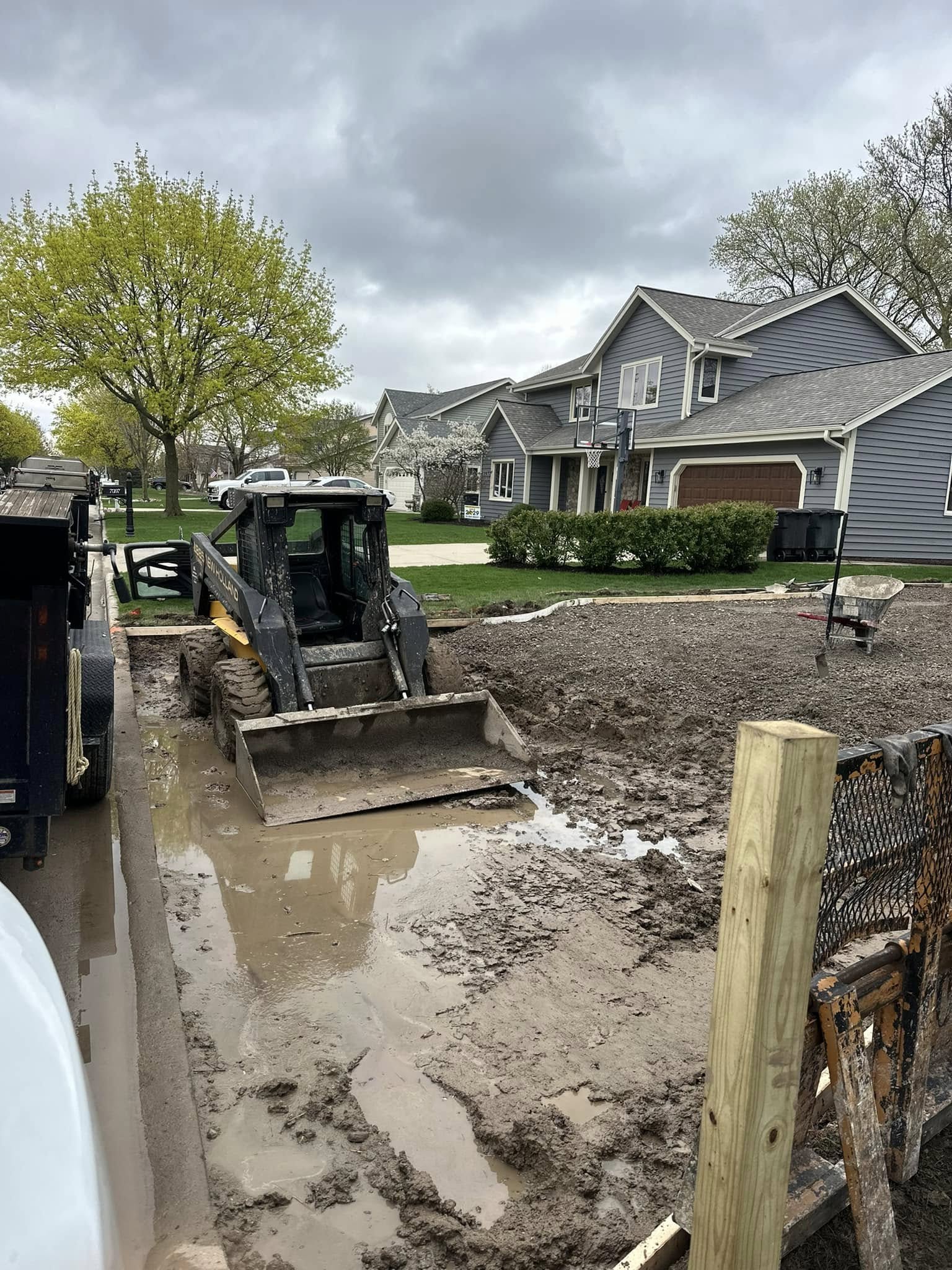 Gravel sub-base fully prepared and formed for concrete pour with workers and equipment staged near garage